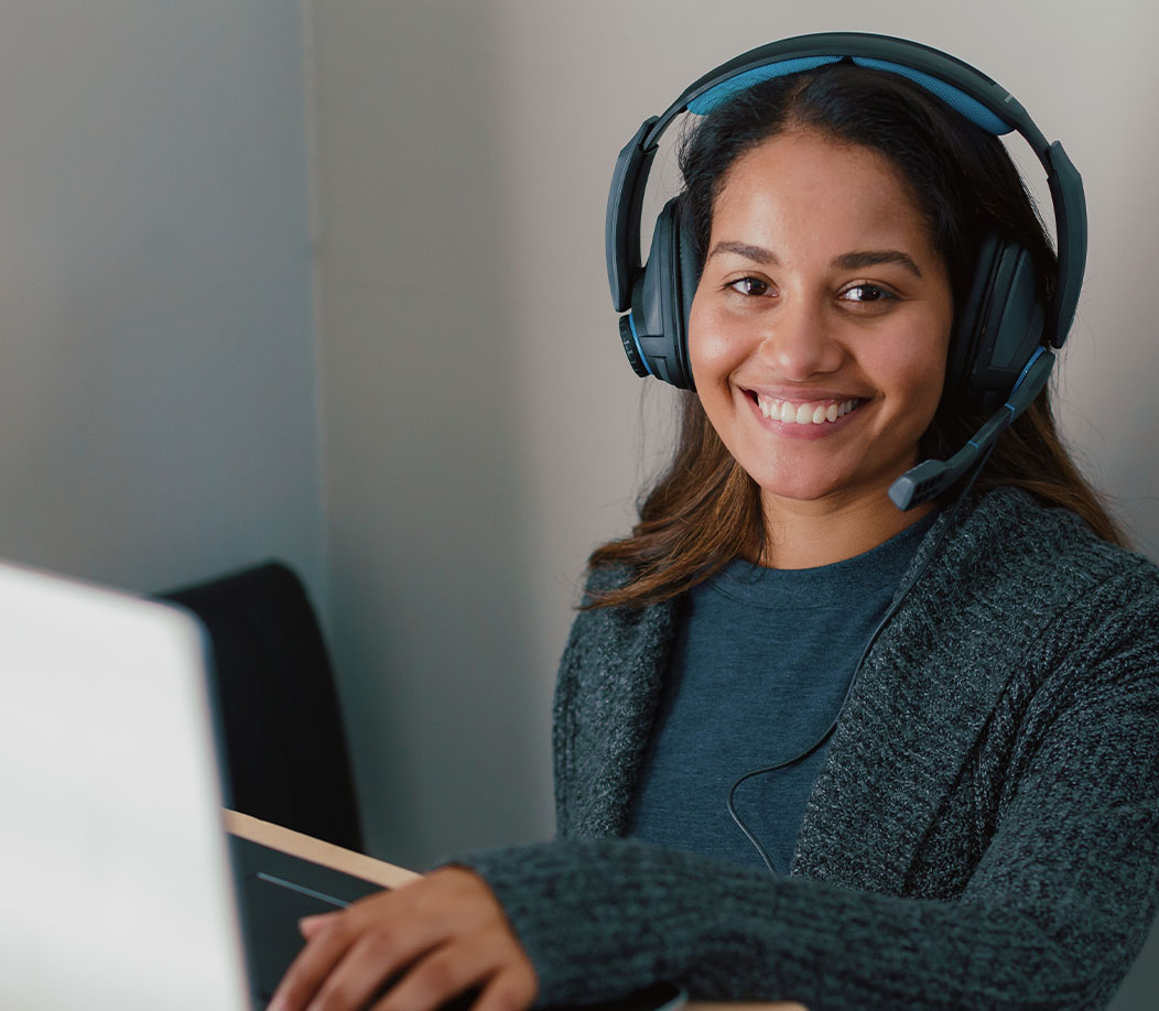 Imagen de persona identificada como mujer en expresión alegre, mirando a la cámara y trabajando con una computadora portátil y auriculares puestos.